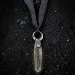 Overhead view of a large, 3-inch clear quartz crystal point topped with a copper electroformed crescent moon holding a shimmering abalone shell inlay. The pendant is suspended from a hand-dyed black silk ribbon with raw edges, laid flat on textured black sand.