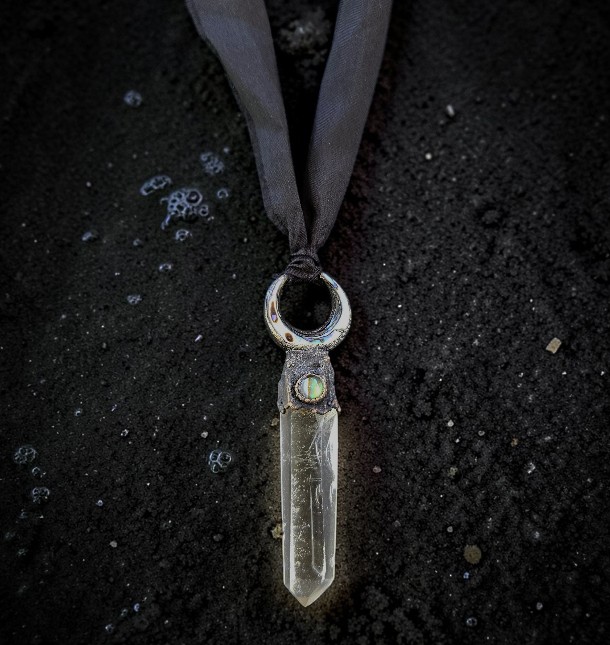Overhead view of a large, 3-inch clear quartz crystal point topped with a copper electroformed crescent moon holding a shimmering abalone shell inlay. The pendant is suspended from a hand-dyed black silk ribbon with raw edges, laid flat on textured black sand.
