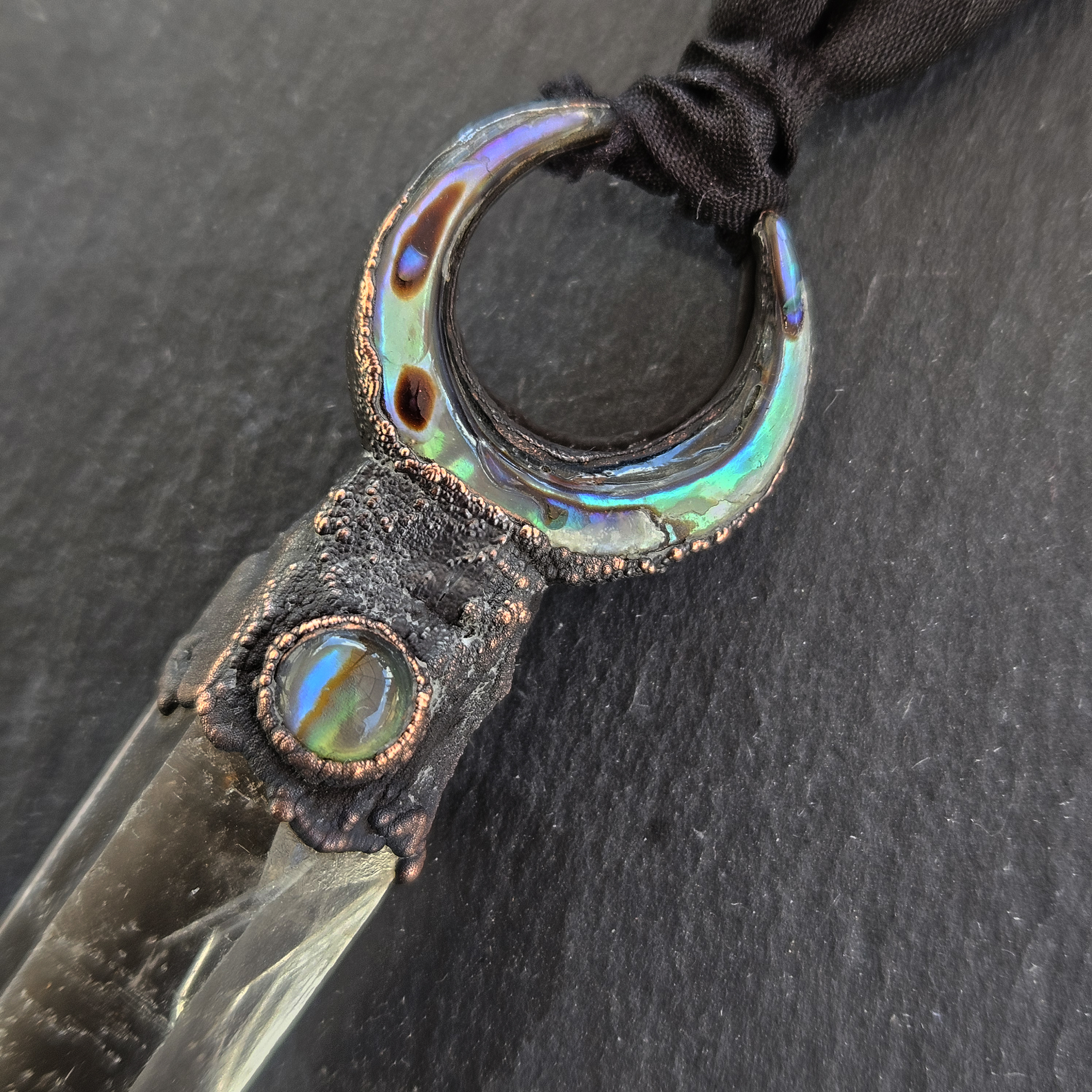 Macro close-up highlighting the vivid green, teal, and purple iridescence of the abalone shell crescent moon and matching round cabochon. The copper setting features a dark, heavily cratered, textured patina.
