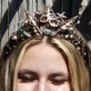 A close-up front view of a blonde model with closed eyes wearing a regal electroformed copper diadem with a high centerpiece peak of sliced sea shells, starfish, pearls, and sea-foam green stones against weathered bamboo backdrop