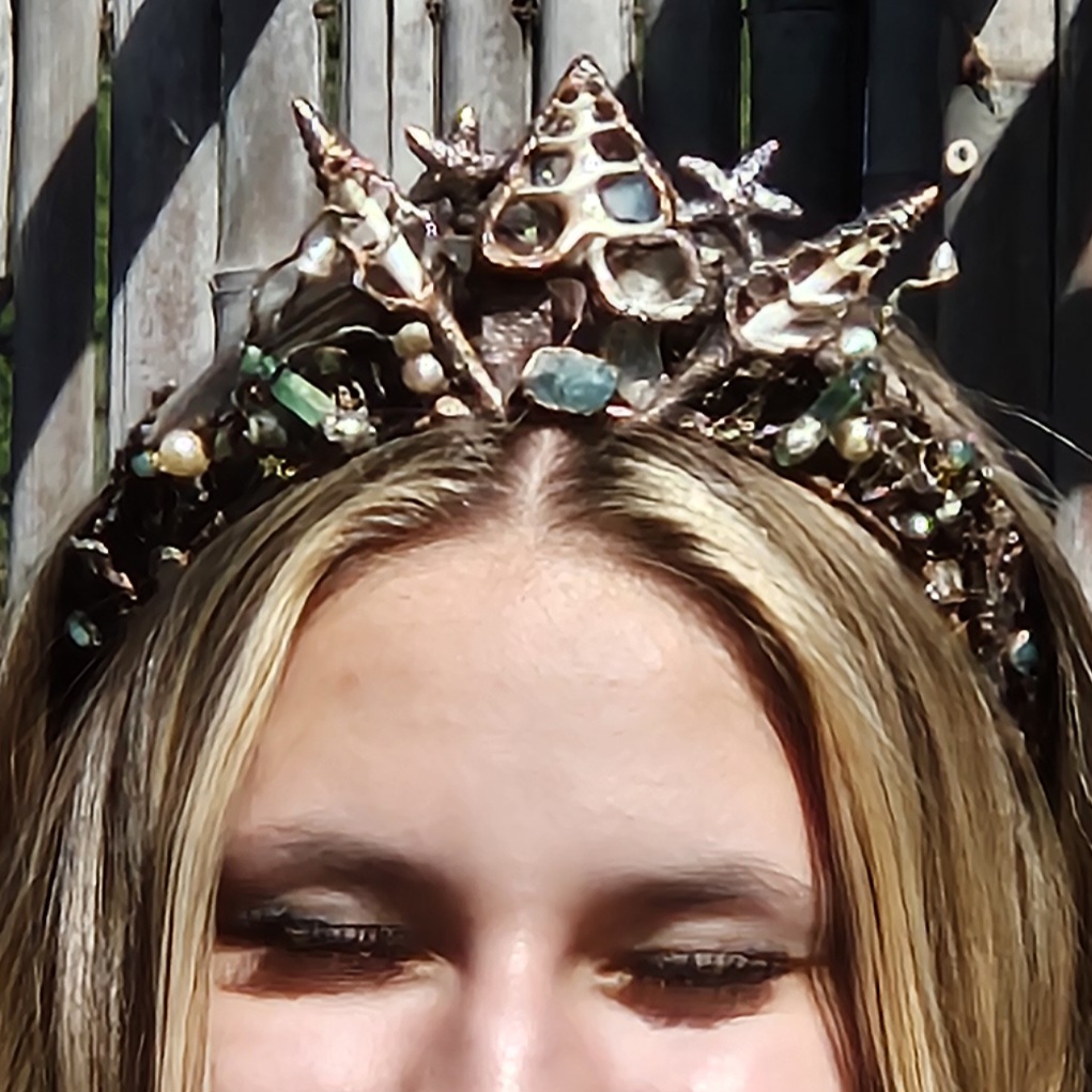 A close-up front view of a blonde model with closed eyes wearing a regal electroformed copper diadem with a high centerpiece peak of sliced sea shells, starfish, pearls, and sea-foam green stones against weathered bamboo backdrop