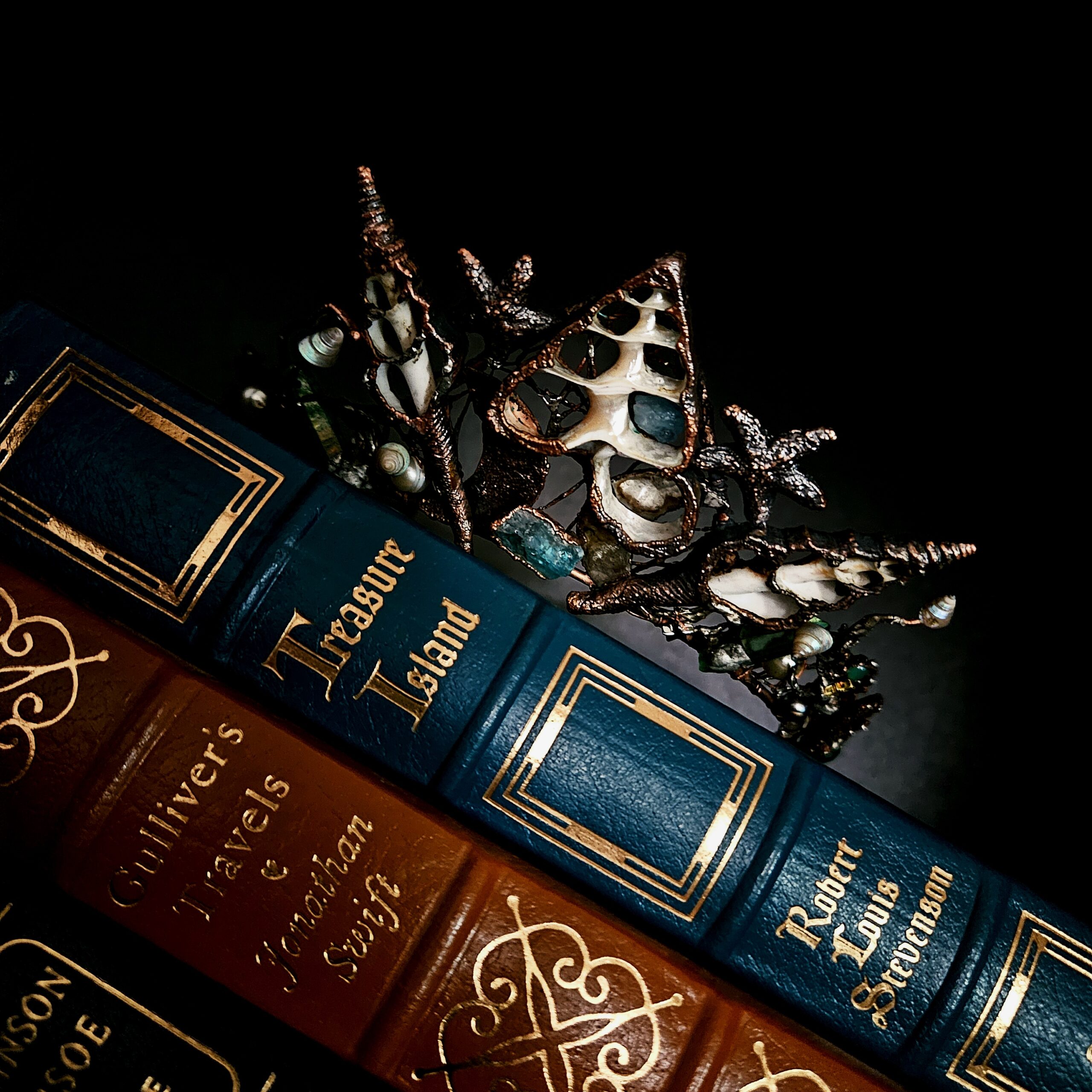 A dark academic close-up of an electroformed copper sea shell crown resting against the gilded leather spines of classic books like Treasure Island inside a moody, shadowed bookcase.