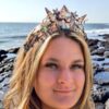 A full-face portrait of a smiling blonde model wearing a handmade electroformed copper sea shell crown, posed against a rugged, rocky coastline with ocean waves crashing in the background under a golden hour sky.