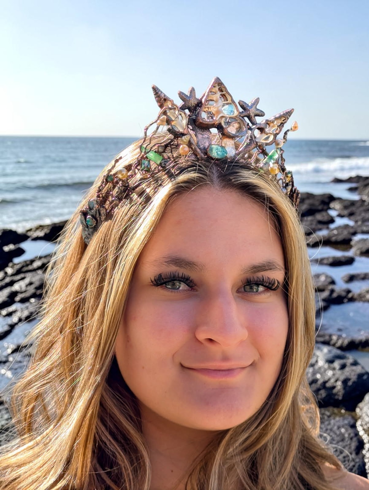 A full-face portrait of a smiling blonde model wearing a handmade electroformed copper sea shell crown, posed against a rugged, rocky coastline with ocean waves crashing in the background under a golden hour sky.