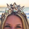 A close-up front view of a blonde model with striking green eyes wearing a regal electroformed copper diadem with a high centerpiece peak of sliced sea shells, starfish, pearls, and sea-foam green stones against a soft, blurred ocean horizon at sunset.