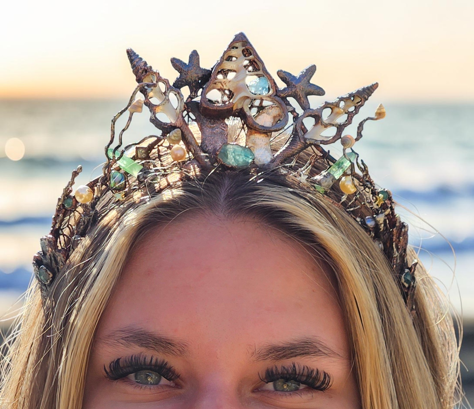 A close-up front view of a blonde model with striking green eyes wearing a regal electroformed copper diadem with a high centerpiece peak of sliced sea shells, starfish, pearls, and sea-foam green stones against a soft, blurred ocean horizon at sunset.