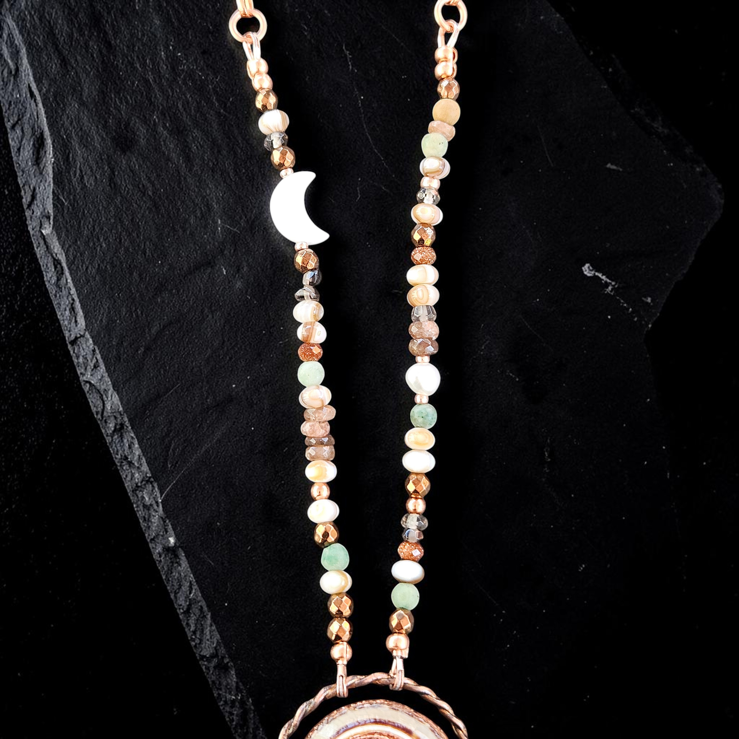 Macro photograph focusing on the asymmetrical bead layout, highlighting the luminous white mother-of-pearl crescent moon charm nested among earth-toned, faceted gemstone beads and metallic accents.