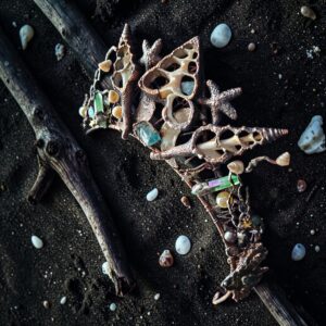 A heavy electroformed copper crown featuring intricate sliced sea shells, starfish, and raw turquoise gemstones, displayed as a flatlay on dark volcanic black sand with weathered driftwood branches.