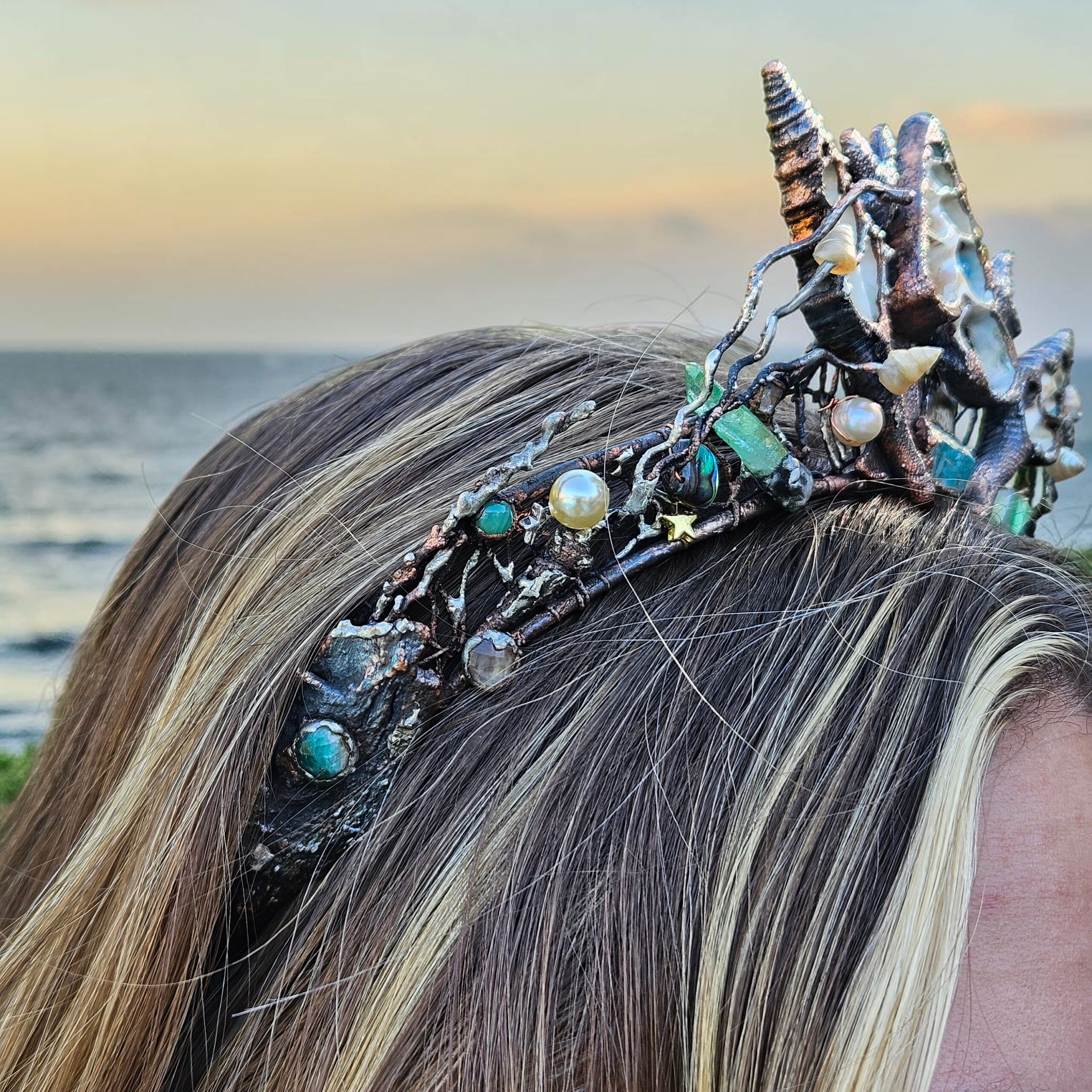 A left profile close-up showing how the textured, barnacle-like electroformed copper band of the crown wraps securely around the side of a blonde model's hair, highlighting embedded high-luster pearls and raw stones.