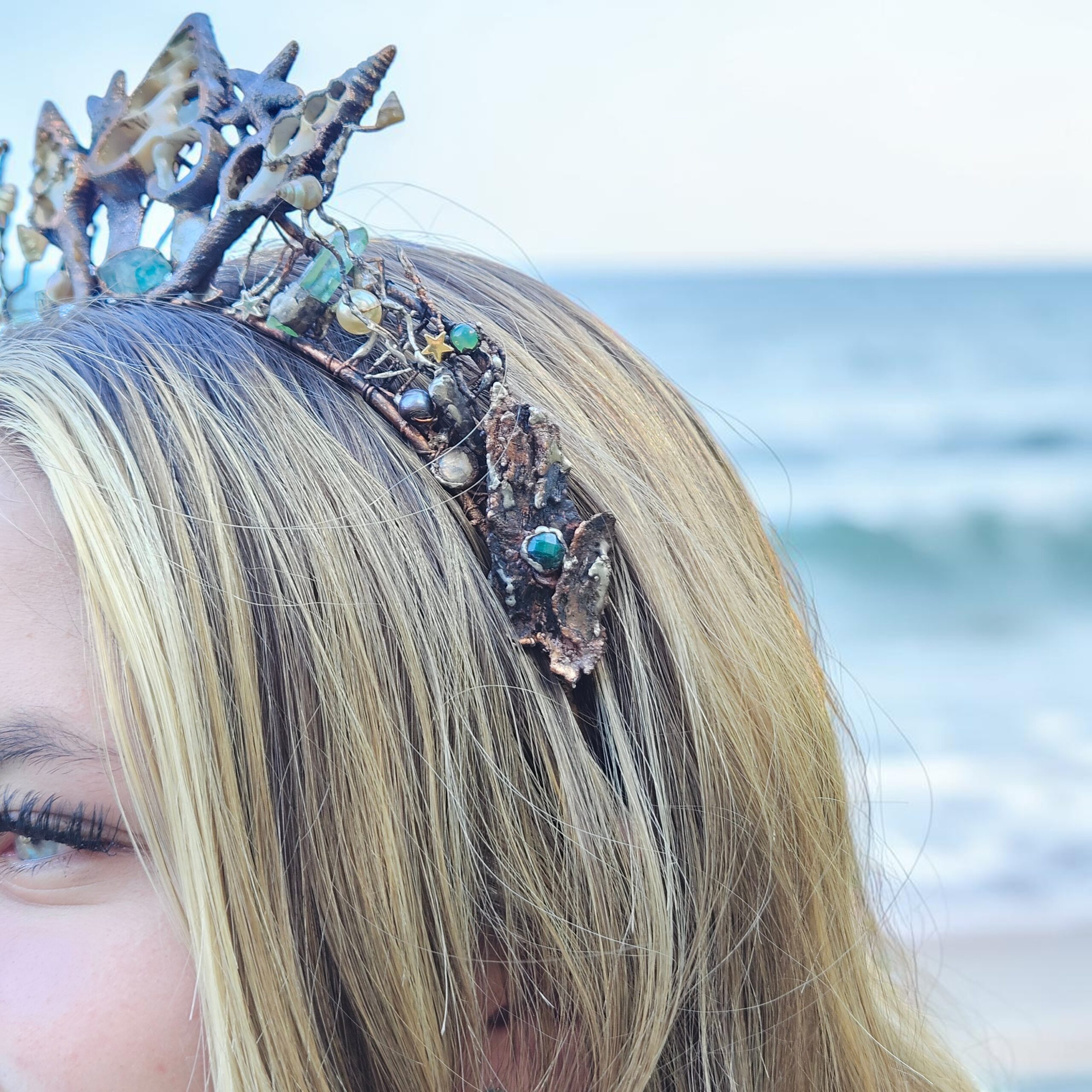 A right profile side view of the sea shell crown, detailing the intricate metallic wirework, small star accents, and heavy organic copper texture against a bright blue ocean background.