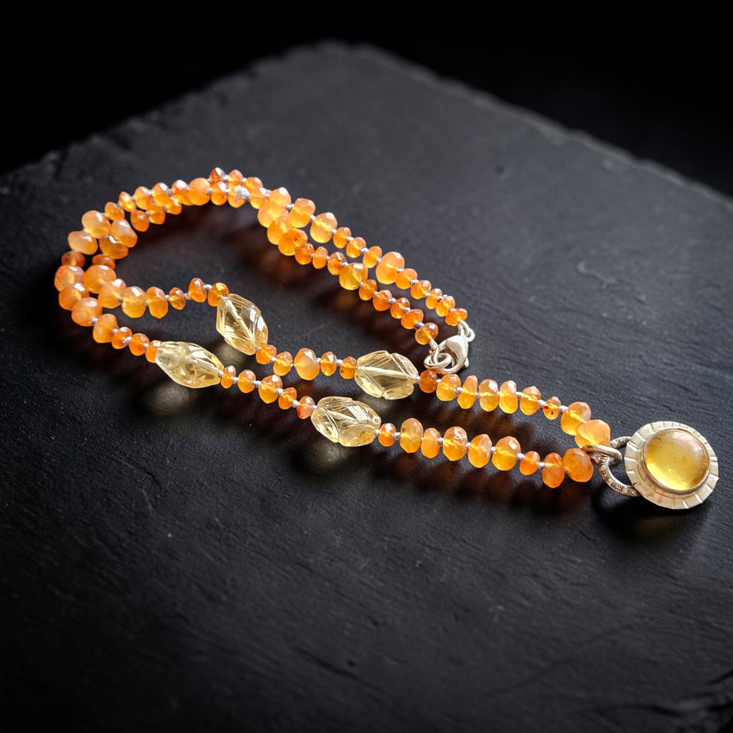 Hand-knotted carnelian bead strand with carved citrine stations and a round amber cabochon sterling silver pendant on a black slate surface.