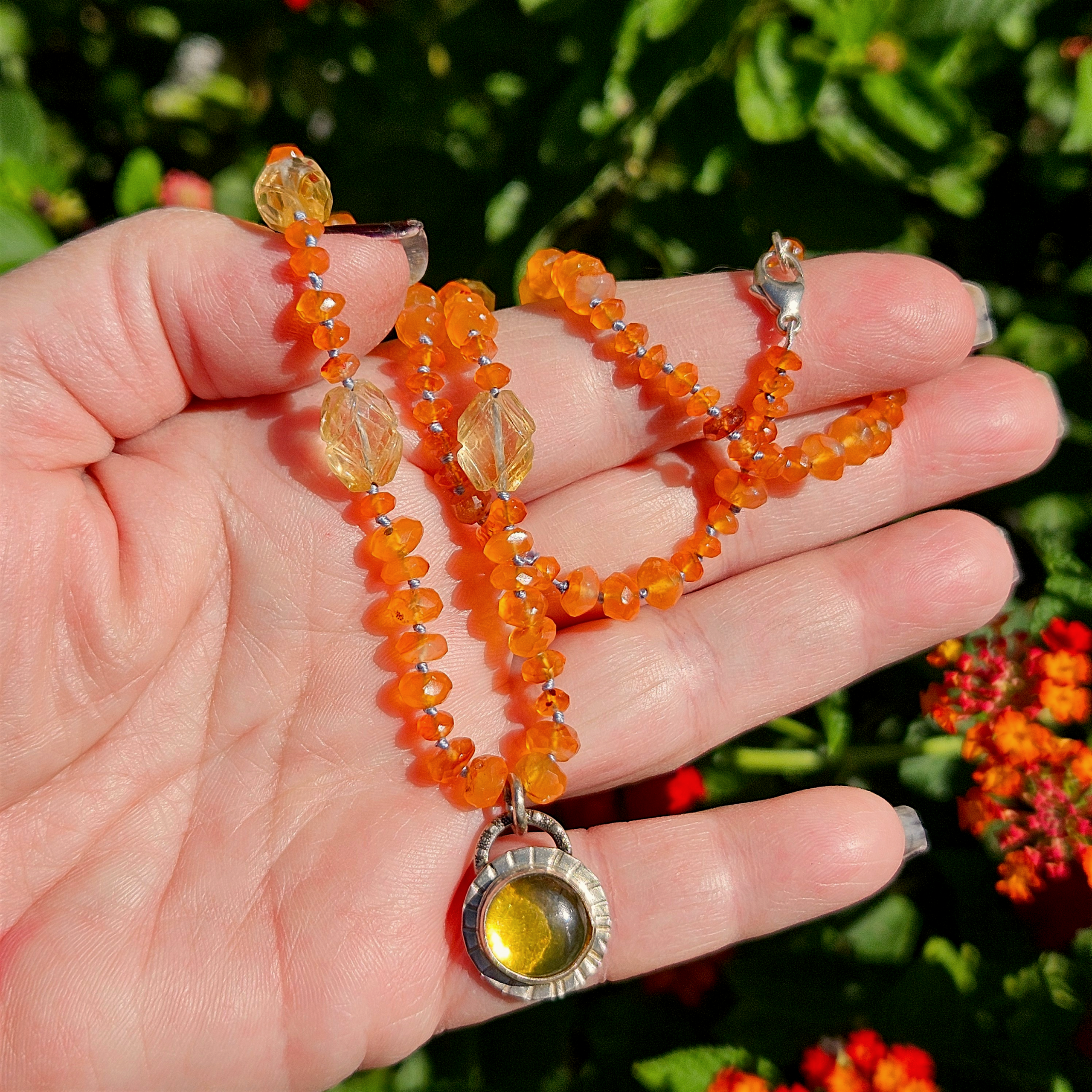 Close-up view of a hand holding the golden amber amulet necklace outdoors in direct sunlight, displaying the radiant warmth of the gems and natural garden background.