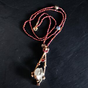Hand-knotted garnet bead necklace with a copper electroformed boxwood branch pendant cradling a clear quartz crystal point and garnet accents on a dark slate background.