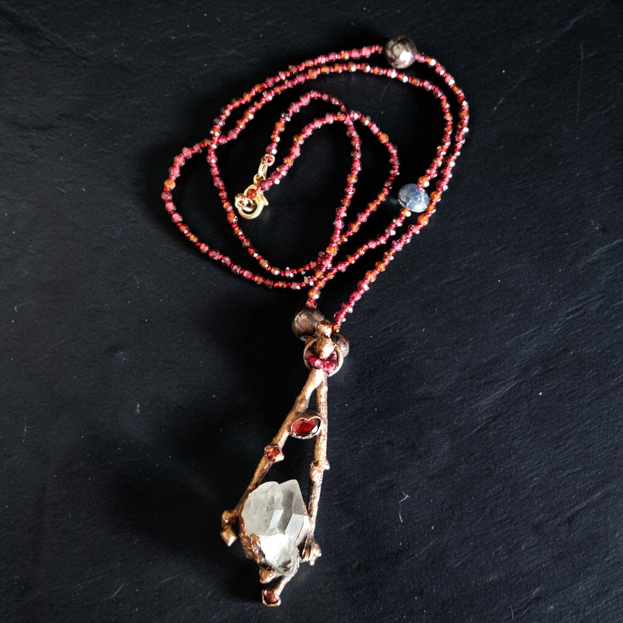 Hand-knotted garnet bead necklace with a copper electroformed boxwood branch pendant cradling a clear quartz crystal point and garnet accents on a dark slate background.