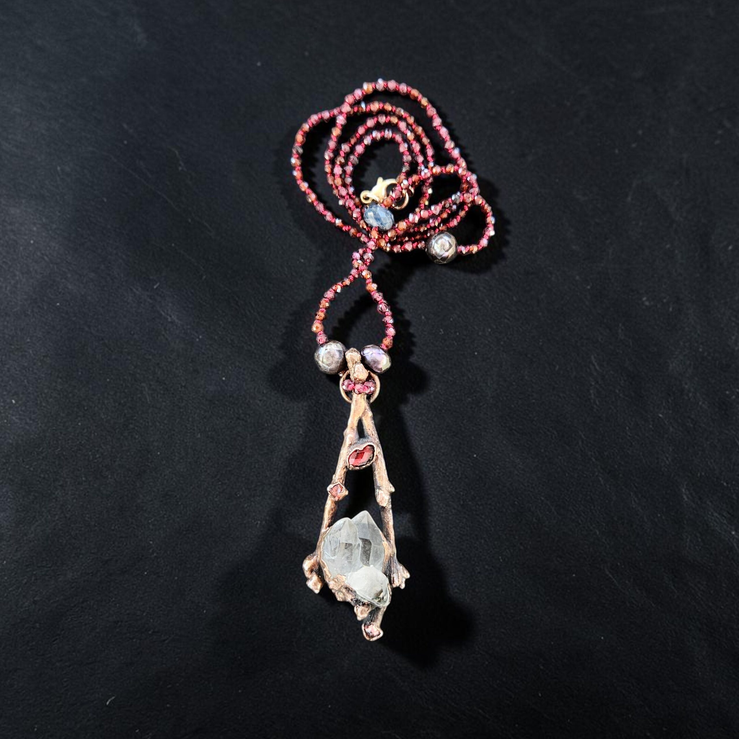 Vertical view of a 24-inch micro-faceted garnet bead strand coiled above a dual-branch copper electroformed pendant with raw clear quartz, dark pearls, and blue sapphire accents.