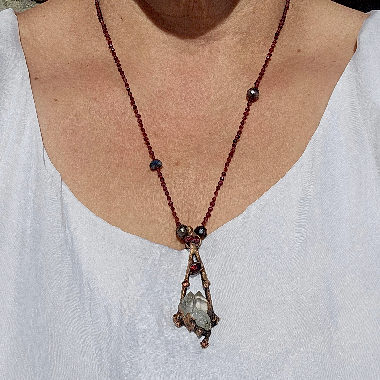 Model wearing the Sticks and Stones garnet necklace with a white dress, showing the 24-inch drop length and how the copper branch pendant sits on the chest.