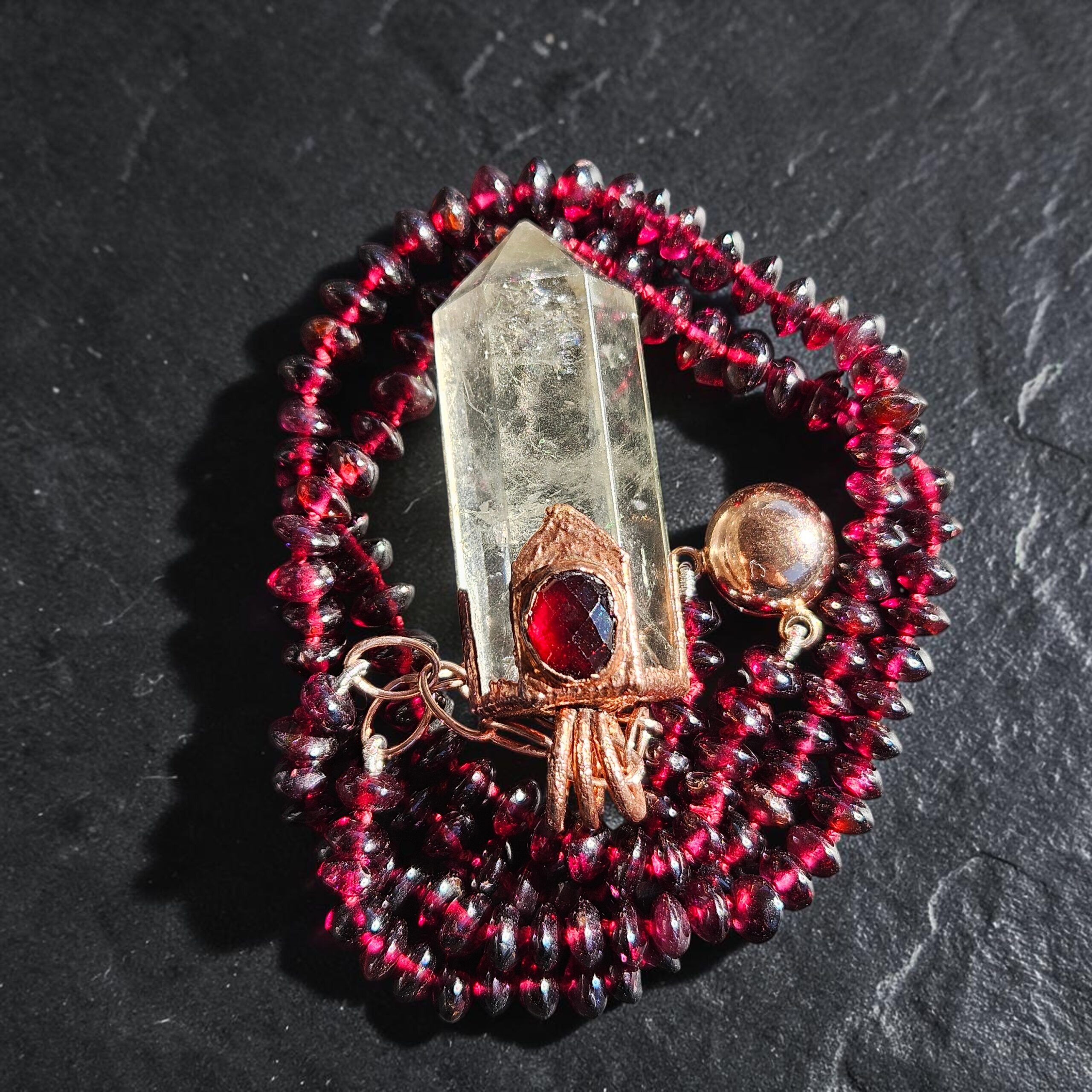 Micro-faceted red garnet bead strand coiled on black slate around a clear quartz point pendant with an embedded faceted garnet in electroformed copper, alongside a rose gold magnetic sphere clasp.