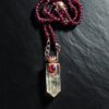 Close-up view of a phantom clear quartz point crowned in copper with a faceted garnet gemstone, hanging from rose gold chain links on a dark slate background.