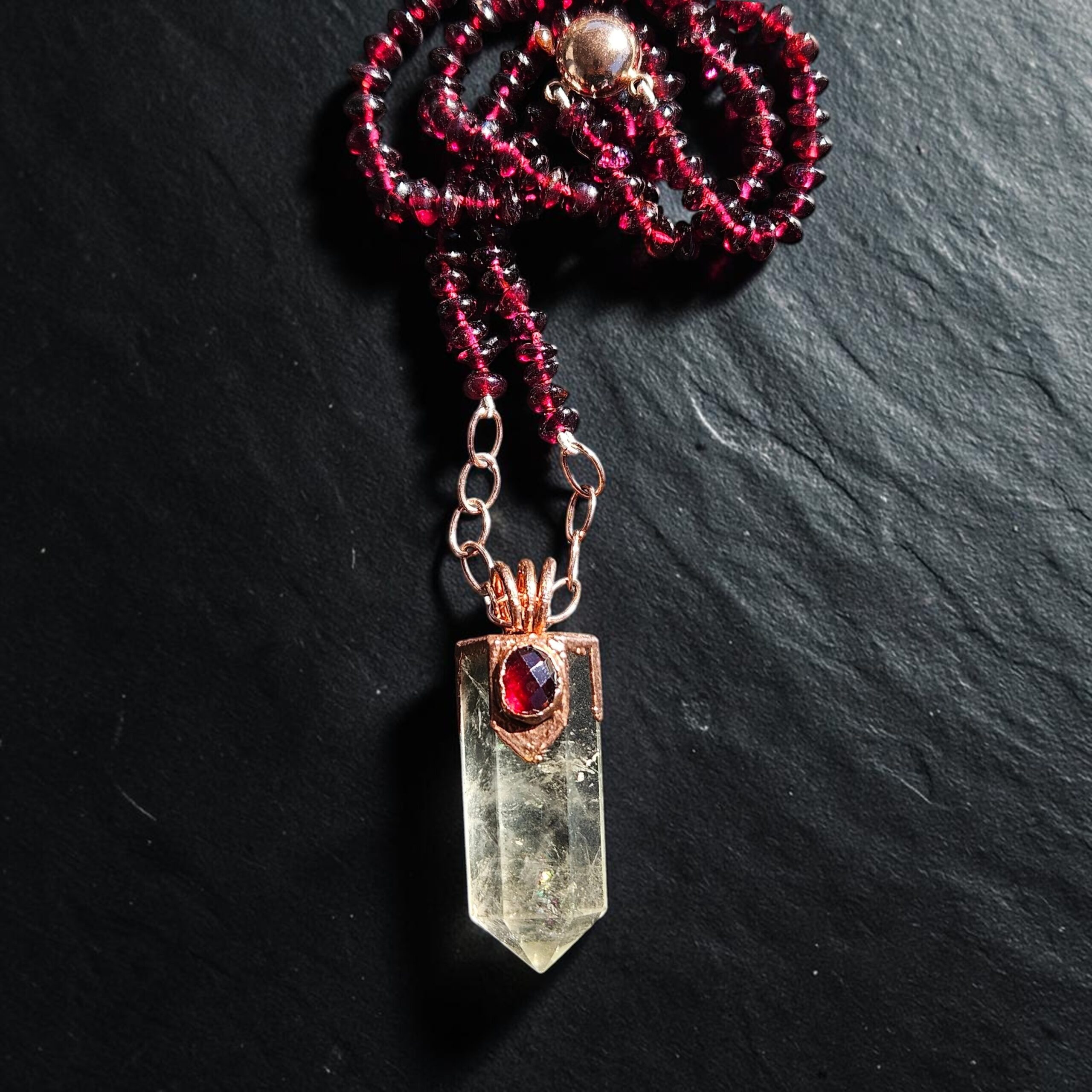 Close-up view of a phantom clear quartz point crowned in copper with a faceted garnet gemstone, hanging from rose gold chain links on a dark slate background.