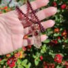 Hand holding the hand-knotted garnet bead necklace outdoors against vibrant garden flowers, displaying the translucent ghost inclusions inside the quartz crystal pendant.