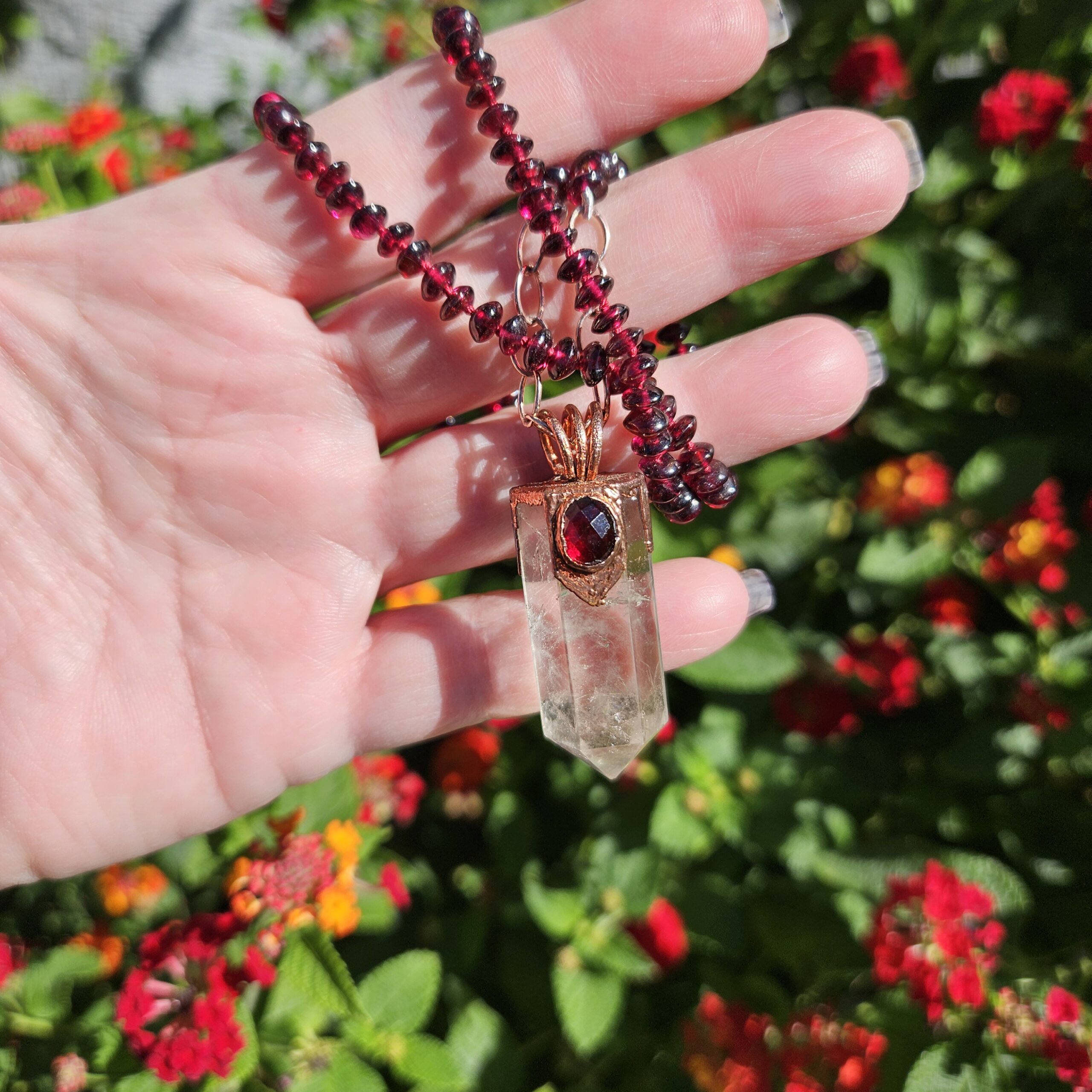 Hand holding the hand-knotted garnet bead necklace outdoors against vibrant garden flowers, displaying the translucent ghost inclusions inside the quartz crystal pendant.