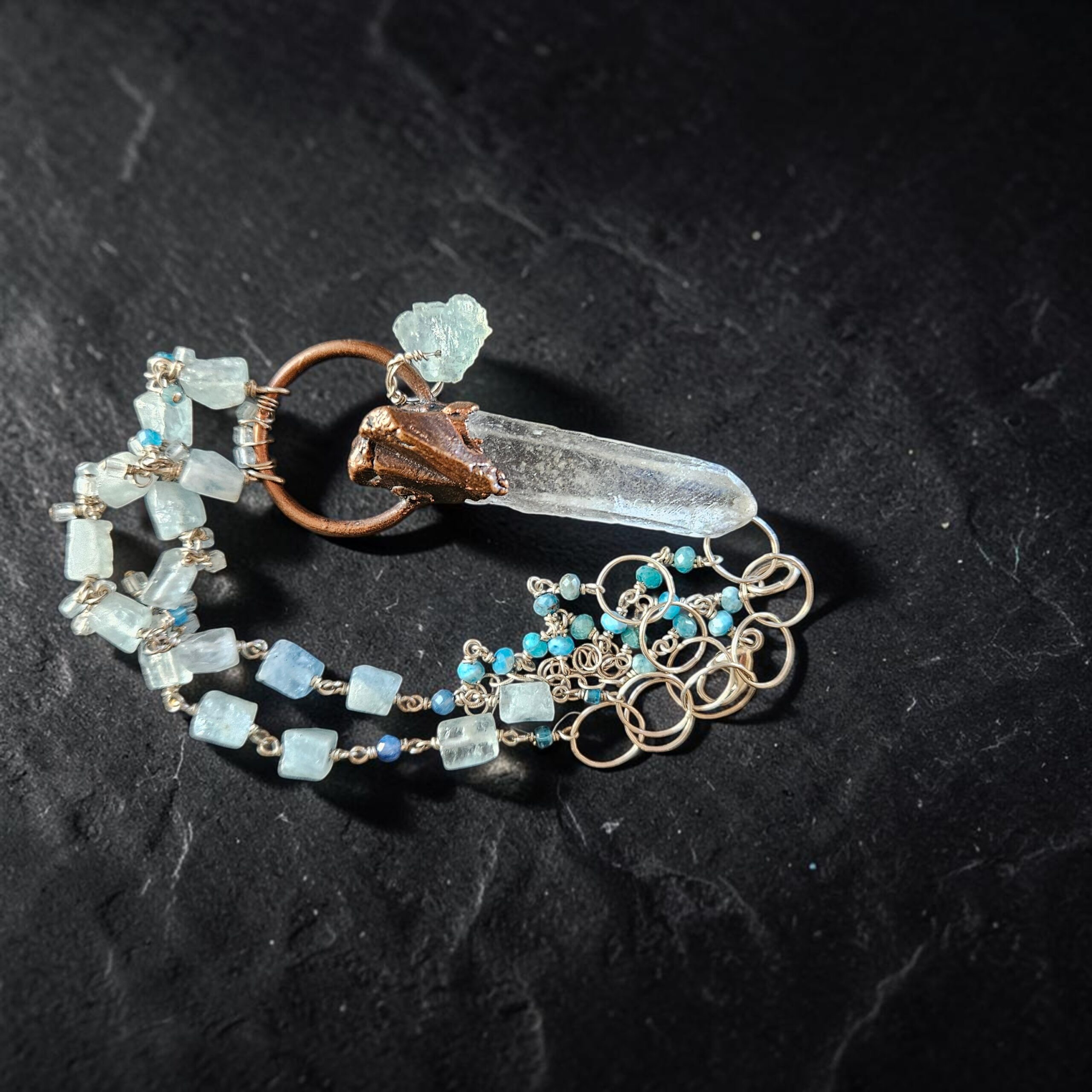 Artistic close-up of the coiled aquamarine and silver chain with the raw clear quartz point pendant resting on black slate under focused light.