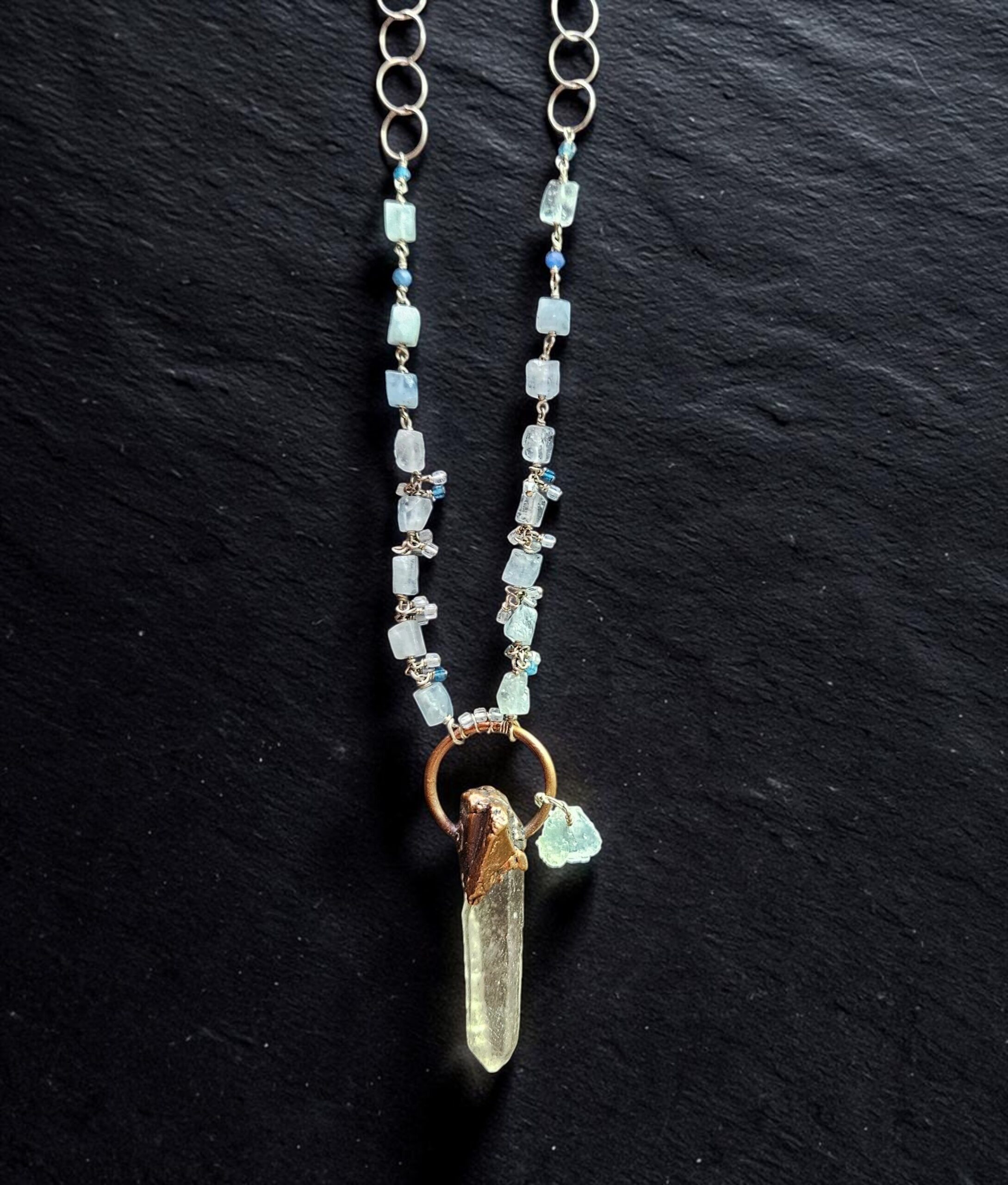 Artistic close-up of the coiled aquamarine and silver chain with the raw clear quartz point pendant resting on black slate under focused light.