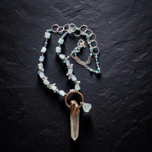 Flat lay view of the Luminous Depths necklace coiled neatly on black slate, highlighting the sterling silver circle links, blue bead details, and raw aquamarine gem strand.