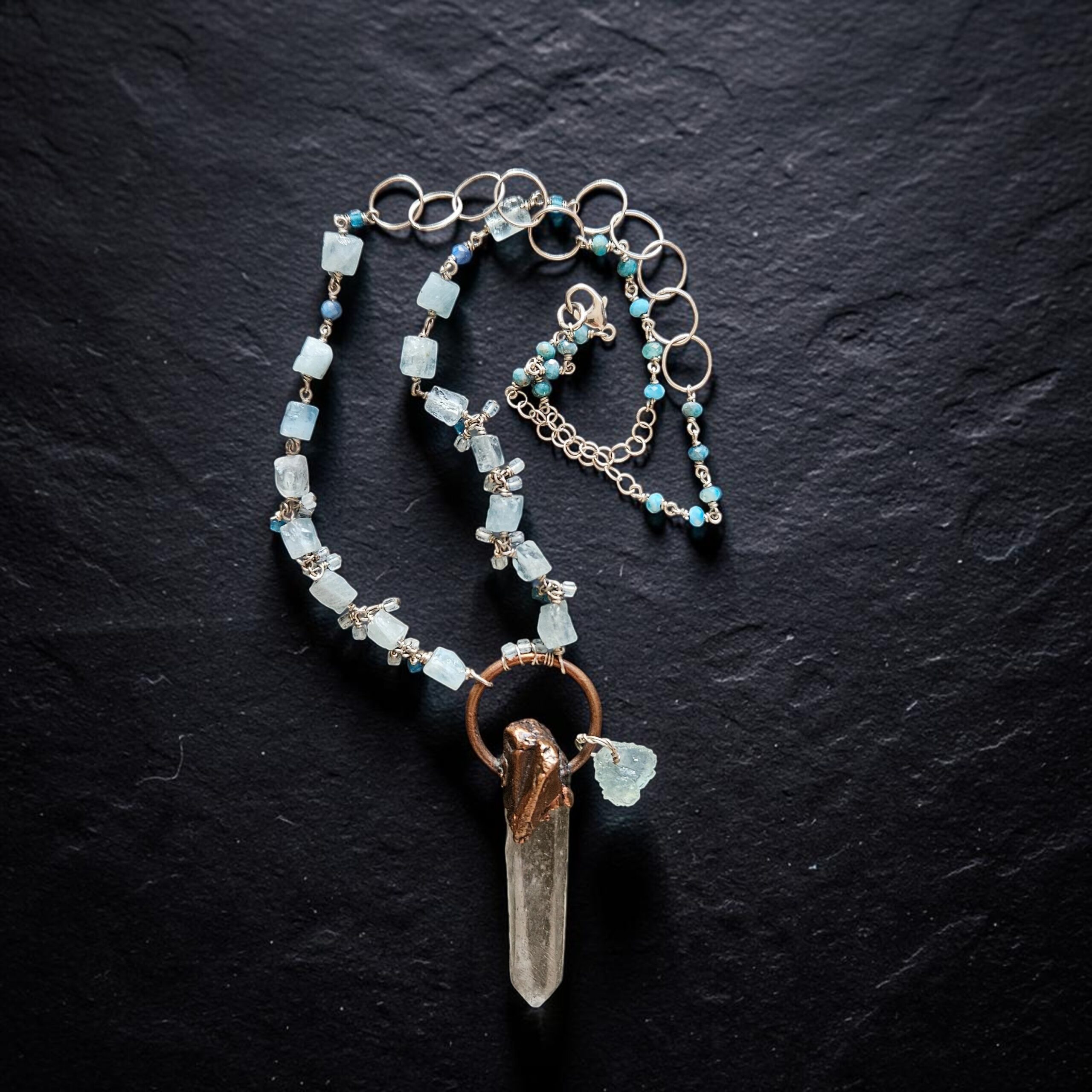 Flat lay view of the Luminous Depths necklace coiled neatly on black slate, highlighting the sterling silver circle links, blue bead details, and raw aquamarine gem strand.