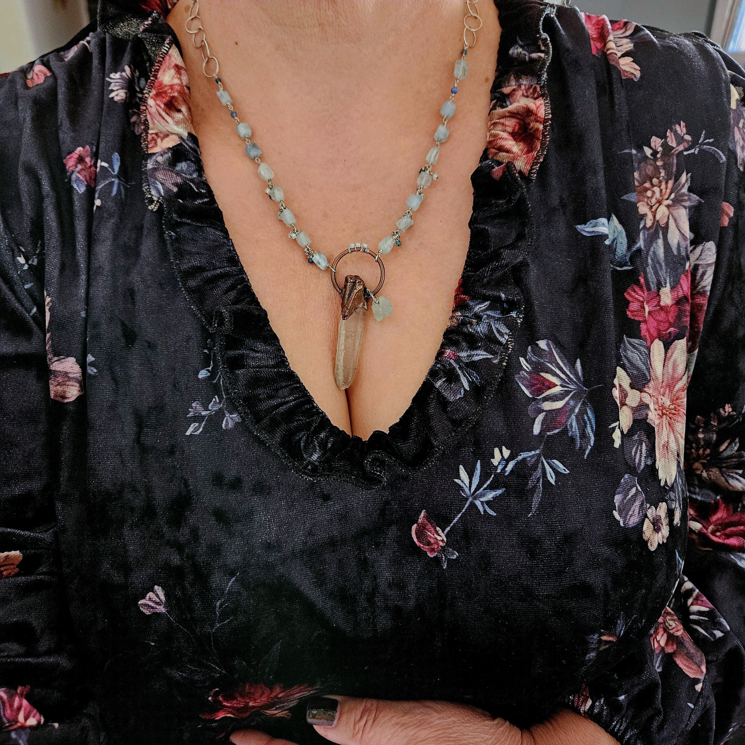 Model wearing the 20-inch Luminous Depths clear quartz and aquamarine pendant necklace over a dark floral velvet top.
