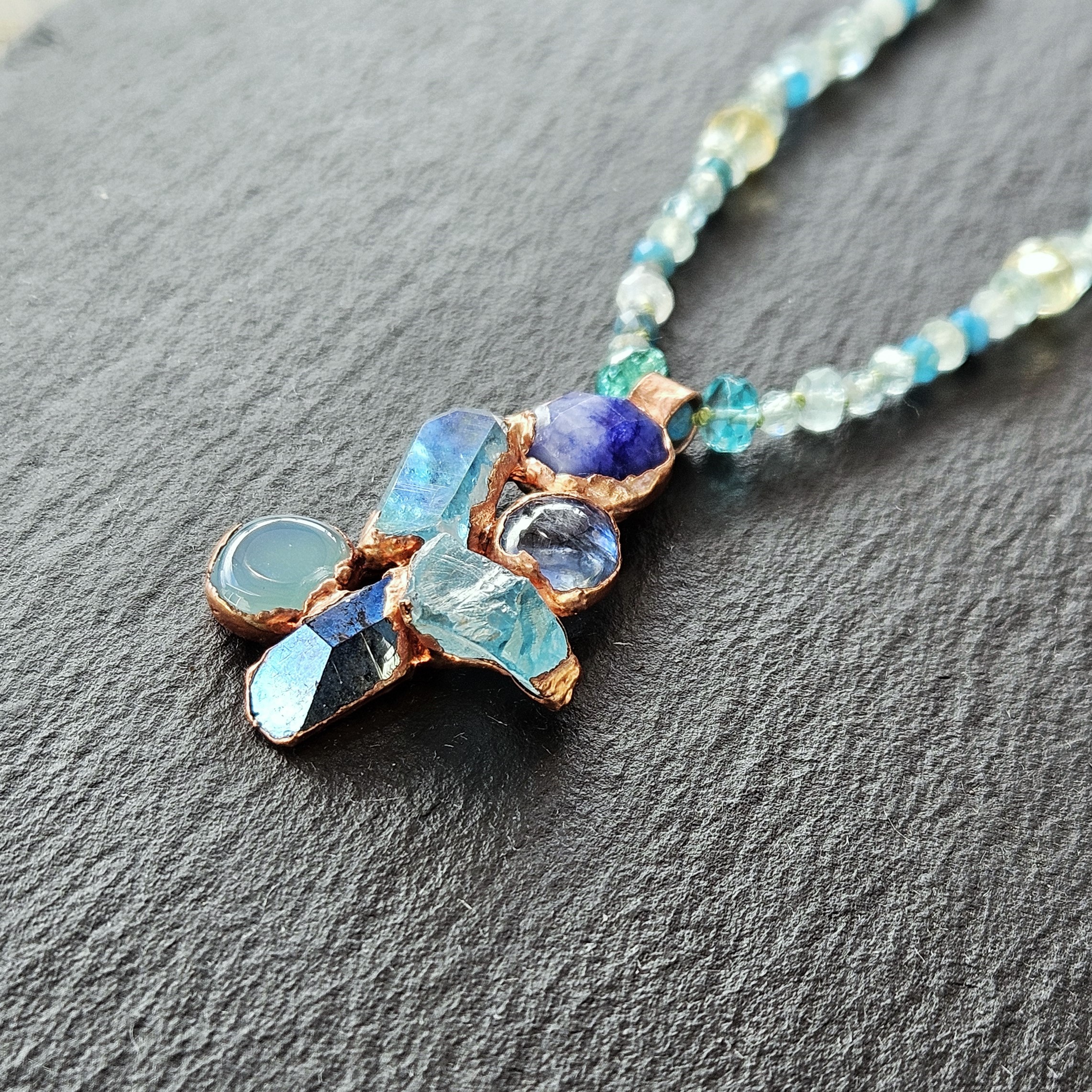 Macro view of the textured copper electroformed crystal cluster pendant lying on dark gray textured slate, capturing the natural facets of raw apatite and kyanite.
