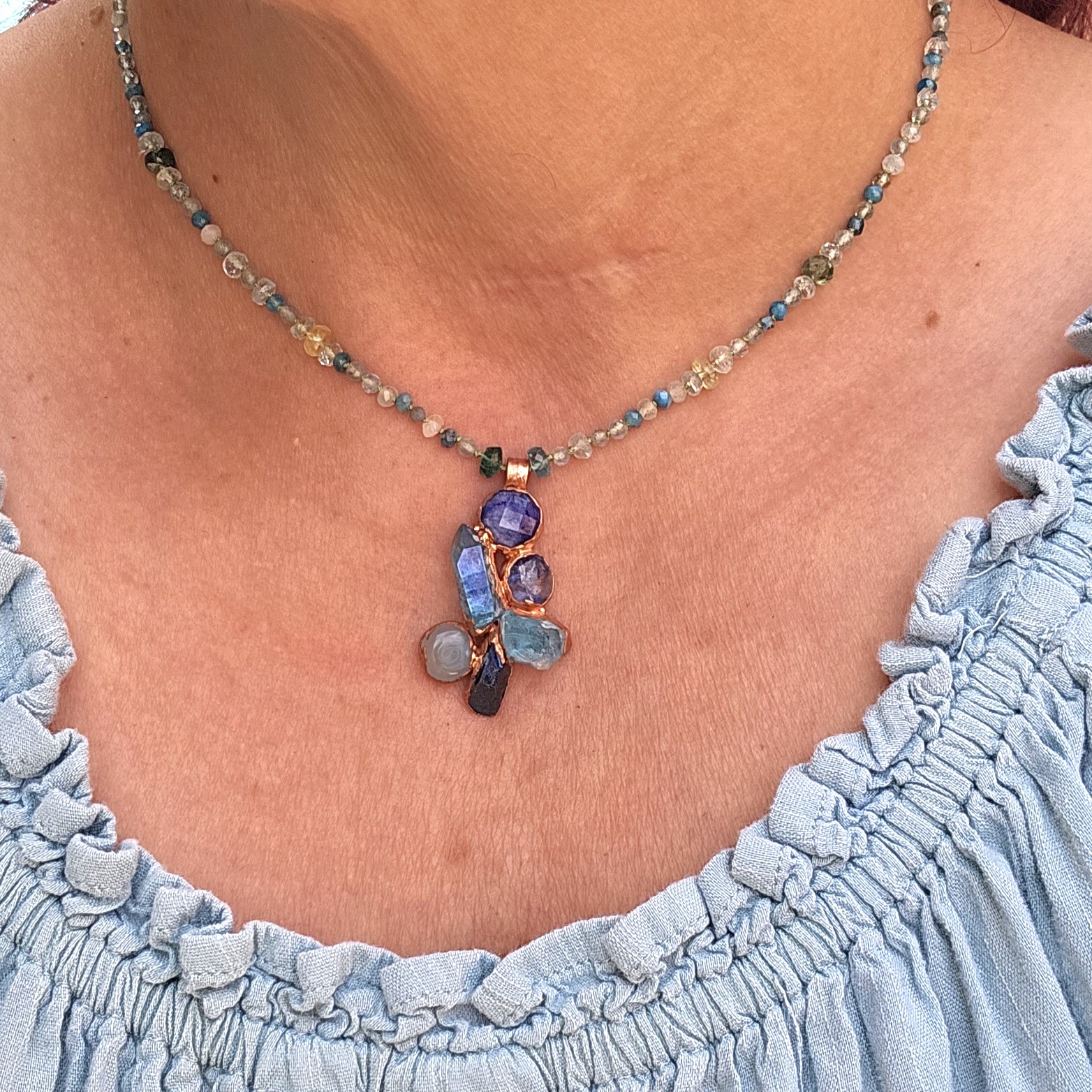 Model wearing the 16.5-inch Sky Realm blue crystal cluster necklace over a light blue ruffled top, showing where the multi-gemstone pendant falls near the collarbone.